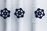 Three pipes with black valve wheels are aligned vertically against a plain background. The pipes and their connecting joints are painted white, contrasting with the dark metal of the valves, which are positioned centrally on each pipe.