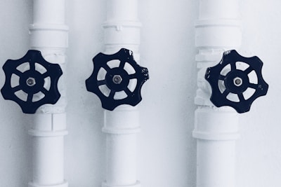 Three pipes with black valve wheels are aligned vertically against a plain background. The pipes and their connecting joints are painted white, contrasting with the dark metal of the valves, which are positioned centrally on each pipe.