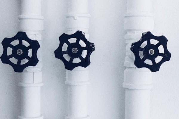 Three pipes with black valve wheels are aligned vertically against a plain background. The pipes and their connecting joints are painted white, contrasting with the dark metal of the valves, which are positioned centrally on each pipe.