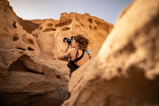 A person is framed against a backdrop of eroded, textured rock formations, capturing the scene with a camera. The setting appears to be a desert with sand-colored cliffs and natural formations.