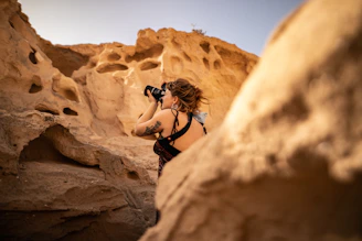 A person is framed against a backdrop of eroded, textured rock formations, capturing the scene with a camera. The setting appears to be a desert with sand-colored cliffs and natural formations.