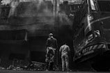 Two individuals stand in front of a large vehicle, possibly a fire truck, near a charred building with smoke billowing from it. The building appears heavily damaged with visible debris on the ground. The image is in black and white, enhancing the dramatic impact.