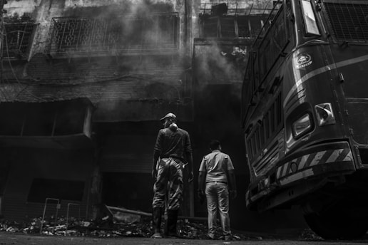 Two individuals stand in front of a large vehicle, possibly a fire truck, near a charred building with smoke billowing from it. The building appears heavily damaged with visible debris on the ground. The image is in black and white, enhancing the dramatic impact.