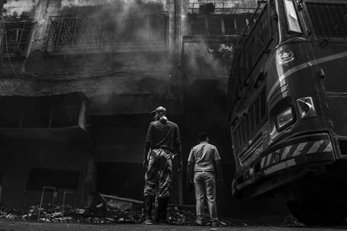 Two individuals stand in front of a large vehicle, possibly a fire truck, near a charred building with smoke billowing from it. The building appears heavily damaged with visible debris on the ground. The image is in black and white, enhancing the dramatic impact.