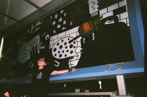 A person stands in front of a large, colorful mural featuring abstract shapes, patterns, and a prominent eye design. The mural incorporates elements such as geometric forms, stars, and a mix of black, white, and vibrant colors. The person has glasses and short hair, appearing to be expressive with open arms.