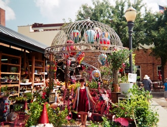 A woman in a red dress stands in front of a garden shop, surrounded by plants and colorful decorations, including balloons inside a decorative metal cage structure. The setting has a vibrant and eclectic array of garden ornaments, with greenery and potted plants adding to the lively ambiance. An outdoor market or urban setting with buildings and trees in the background completes the scene.