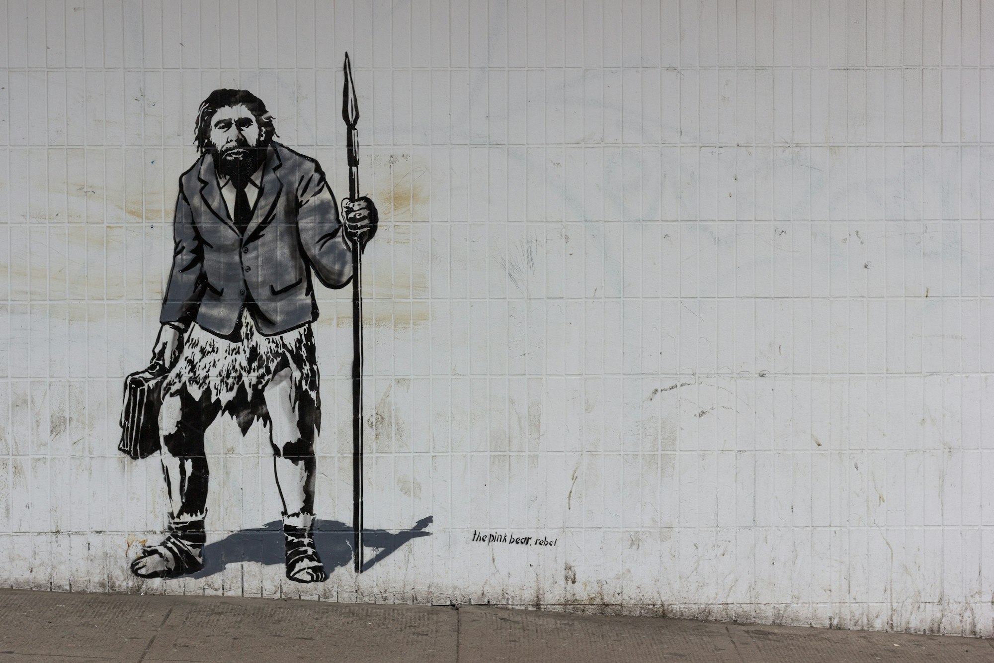 Lovely stencil work by thepinkbear.rebel street artist of a neanderthal man holding a briefcase in one hand and a spear in the other. Wearing a shirt, tie and suit jacket over an animal skin. Ready for a solid days work.