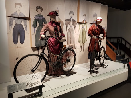 Two mannequins dressed in historical cycling attire stand beside period bicycles. The setting appears to be a museum exhibit with fashion sketches in the background, showcasing clothing styles from a bygone era.