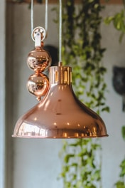 A copper pendant light fixture with a glossy finish is suspended from a metal chain. The fixture features spherical decorative elements and has a modern, industrial design. In the background, there is soft-focus greenery, possibly indoor plants, adding a natural touch to the setting.