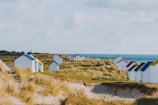 A series of small, white beach huts with colorful roofs are scattered along a sandy coastal area with dunes and grass. The sea is visible in the background with a clear blue sky overhead.