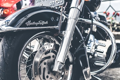Close-up of a motorcycle focusing on the front wheel and fork. The shiny metal parts and intricate spokes are highlighted, with the bike's name 'Heritage Softail' visible on the fender. The background includes blurred elements like another vehicle and flowers.