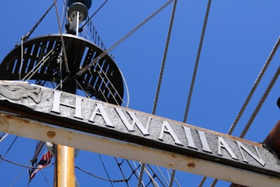 A view of a ship's mast against a clear blue sky, featuring a wooden sign engraved with 'HAWAIIAN' and decorated with a carved emblem. Several ropes and rigging elements are visible, leading toward the crow's nest.