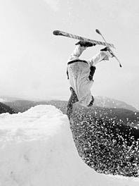 A snowboarder is performing an inverted aerial trick off a snow ramp, with snow spraying in the air. The scene captures a sense of motion and skill as the athlete executes a flip, surrounded by a snowy landscape.
