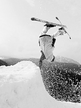 A snowboarder is performing an inverted aerial trick off a snow ramp, with snow spraying in the air. The scene captures a sense of motion and skill as the athlete executes a flip, surrounded by a snowy landscape.