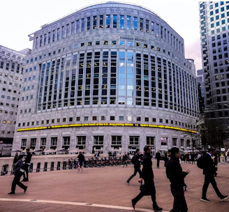 A large, modern office building with reflective glass and a prominent logo at the top. There are people walking in the foreground, suggesting a business district atmosphere. The architecture is imposing, with evenly spaced windows and a ticker displaying news headlines across the front of the building.
