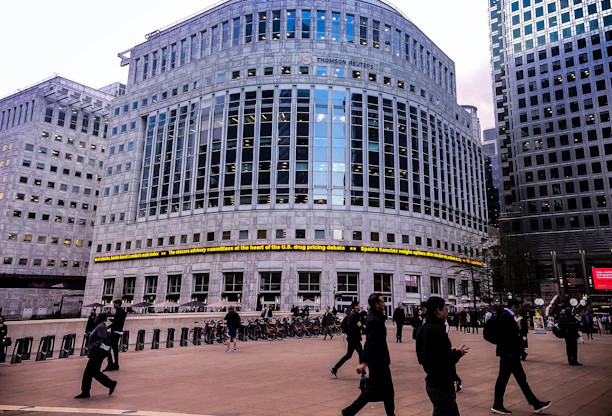 A large, modern office building with reflective glass and a prominent logo at the top. There are people walking in the foreground, suggesting a business district atmosphere. The architecture is imposing, with evenly spaced windows and a ticker displaying news headlines across the front of the building.