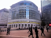 A large, modern office building with reflective glass and a prominent logo at the top. There are people walking in the foreground, suggesting a business district atmosphere. The architecture is imposing, with evenly spaced windows and a ticker displaying news headlines across the front of the building.
