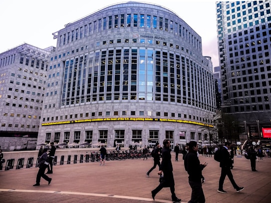 A large, modern office building with reflective glass and a prominent logo at the top. There are people walking in the foreground, suggesting a business district atmosphere. The architecture is imposing, with evenly spaced windows and a ticker displaying news headlines across the front of the building.