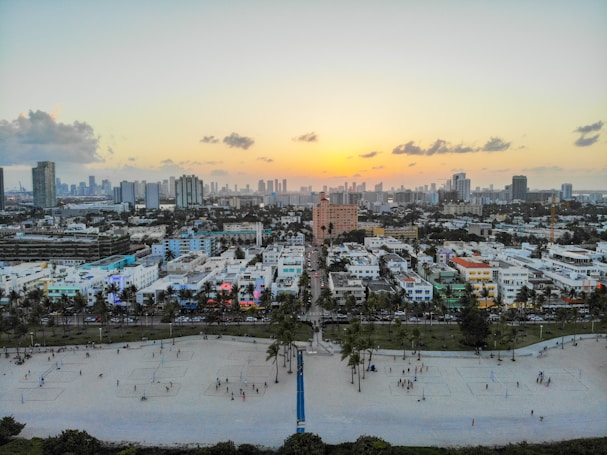 A vibrant cityscape during sunset features a sprawling urban area with numerous high-rise buildings in the background and colorful low-rise structures in the foreground. A beach area with sand volleyball courts and scattered people is visible at the bottom, leading to a line of palm trees and a busy street running parallel to the shore.