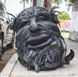 A large sculpture of a human face has been created from recycled black tires. The detailed features, such as eyes and mouth, are prominently carved, and the sculpture sits on a sidewalk near a parked car. In the background, there are colorful murals on the walls and some greenery present.