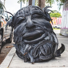 A large sculpture of a human face has been created from recycled black tires. The detailed features, such as eyes and mouth, are prominently carved, and the sculpture sits on a sidewalk near a parked car. In the background, there are colorful murals on the walls and some greenery present.