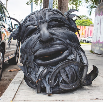 A large sculpture of a human face has been created from recycled black tires. The detailed features, such as eyes and mouth, are prominently carved, and the sculpture sits on a sidewalk near a parked car. In the background, there are colorful murals on the walls and some greenery present.