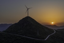 A wind turbine is situated atop a lush, green hill with a winding road leading up to it. A setting sun creates a warm, golden glow in the sky, casting shadows over the mountainous landscape.