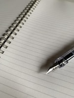 A lined notebook with a spiral binding running along the left side. A fountain pen rests on the lower right corner of the notebook, with a clear body and a shiny nib.