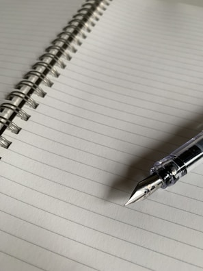 A lined notebook with a spiral binding running along the left side. A fountain pen rests on the lower right corner of the notebook, with a clear body and a shiny nib.