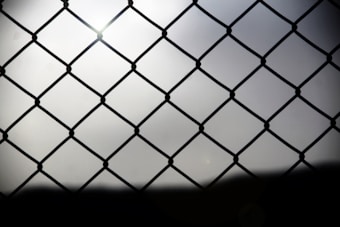 A chain-link fence is shown in silhouette against a bright, diffused light background. The fence creates a repeating diamond pattern with thin wires connected at nodes. The background appears to be overcast, soft, and slightly hazy, adding a feeling of depth.