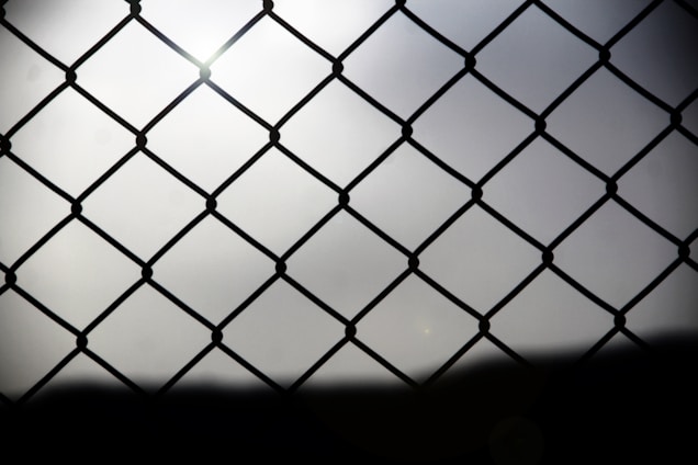 A chain-link fence is shown in silhouette against a bright, diffused light background. The fence creates a repeating diamond pattern with thin wires connected at nodes. The background appears to be overcast, soft, and slightly hazy, adding a feeling of depth.