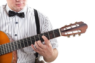 A person wearing a striped shirt with suspenders is playing a classical guitar. The focus is on the neck of the guitar and the person's hand positioning on the strings. The person's face is partially visible, and they are wearing a bow tie.