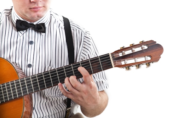 A person wearing a striped shirt with suspenders is playing a classical guitar. The focus is on the neck of the guitar and the person's hand positioning on the strings. The person's face is partially visible, and they are wearing a bow tie.
