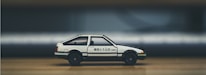 A white and black toy car with Japanese characters on the side is placed on a wooden surface. The background is blurred, focusing attention on the car model, which resembles a classic sports car design with compact dimensions.