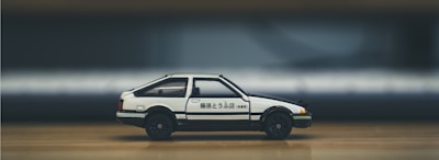 A white and black toy car with Japanese characters on the side is placed on a wooden surface. The background is blurred, focusing attention on the car model, which resembles a classic sports car design with compact dimensions.