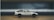 A white and black toy car with Japanese characters on the side is placed on a wooden surface. The background is blurred, focusing attention on the car model, which resembles a classic sports car design with compact dimensions.