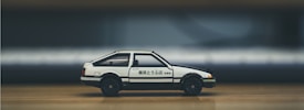 A white and black toy car with Japanese characters on the side is placed on a wooden surface. The background is blurred, focusing attention on the car model, which resembles a classic sports car design with compact dimensions.