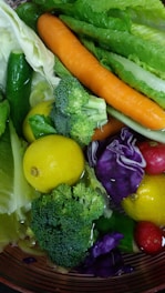 A colorful assortment of fresh vegetables and fruits including a carrot, lemons, broccoli, leafy greens, cucumber, red radishes, and purple cabbage. They are closely packed together, reflecting various shades and textures under a light source, with some water droplets visible on their surfaces.