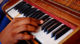 A hand playing an antique harmonium with black and white keys and a wooden body. The focus is on the fingers pressing the keys, capturing the texture and warmth of the instrument.