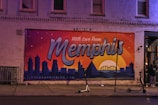 A colorful mural is painted on a brick building, featuring a sunset view of a city's skyline with the text 'With Love From Memphis'. There are scooters parked on the sidewalk and a few individuals standing near the building's entrance. The mural includes a depiction of the Hernando de Soto Bridge.