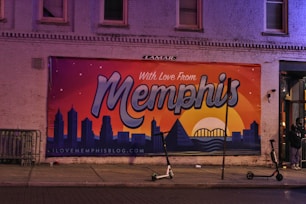 A colorful mural is painted on a brick building, featuring a sunset view of a city's skyline with the text 'With Love From Memphis'. There are scooters parked on the sidewalk and a few individuals standing near the building's entrance. The mural includes a depiction of the Hernando de Soto Bridge.