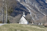 Nestled in a grassy landscape, a quaint white church with a pointed spire stands surrounded by leafless trees. A stone wall borders the rolling grassy area. In the background, a rugged mountain slope dominates the scene under a clear blue sky.