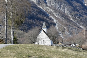Nestled in a grassy landscape, a quaint white church with a pointed spire stands surrounded by leafless trees. A stone wall borders the rolling grassy area. In the background, a rugged mountain slope dominates the scene under a clear blue sky.