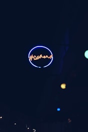 A neon sign with a circular outline emitting a blue glow, featuring red neon text in an urban setting. Additional small colored light spots are visible in the background.