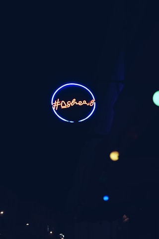 A neon sign with a circular outline emitting a blue glow, featuring red neon text in an urban setting. Additional small colored light spots are visible in the background.