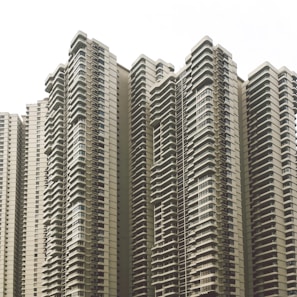 A cluster of modern high-rise apartment buildings with numerous balconies and uniform architectural design. The buildings feature a neutral color scheme and numerous windows, creating a repetitive and geometric pattern.