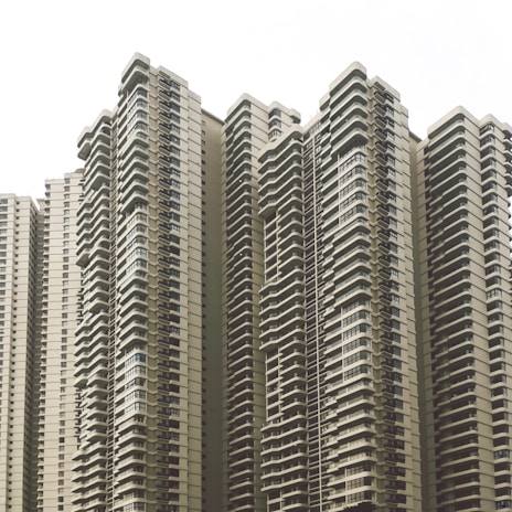 A cluster of modern high-rise apartment buildings with numerous balconies and uniform architectural design. The buildings feature a neutral color scheme and numerous windows, creating a repetitive and geometric pattern.