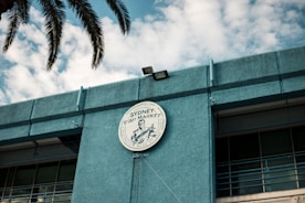 A circular sign for the Sydney Fish Market is prominently displayed on the exterior of a blue building. The sign features a graphic and writing, with palm leaves partially visible above. The sky in the background is filled with scattered clouds.