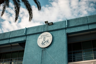 A circular sign for the Sydney Fish Market is prominently displayed on the exterior of a blue building. The sign features a graphic and writing, with palm leaves partially visible above. The sky in the background is filled with scattered clouds.