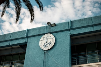 A circular sign for the Sydney Fish Market is prominently displayed on the exterior of a blue building. The sign features a graphic and writing, with palm leaves partially visible above. The sky in the background is filled with scattered clouds.